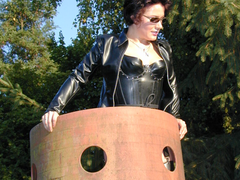 Frau-mit-Sonnenbrille-in-Lack-und-Leder-posiert-draussen-auf-einer-Metallskulptur
